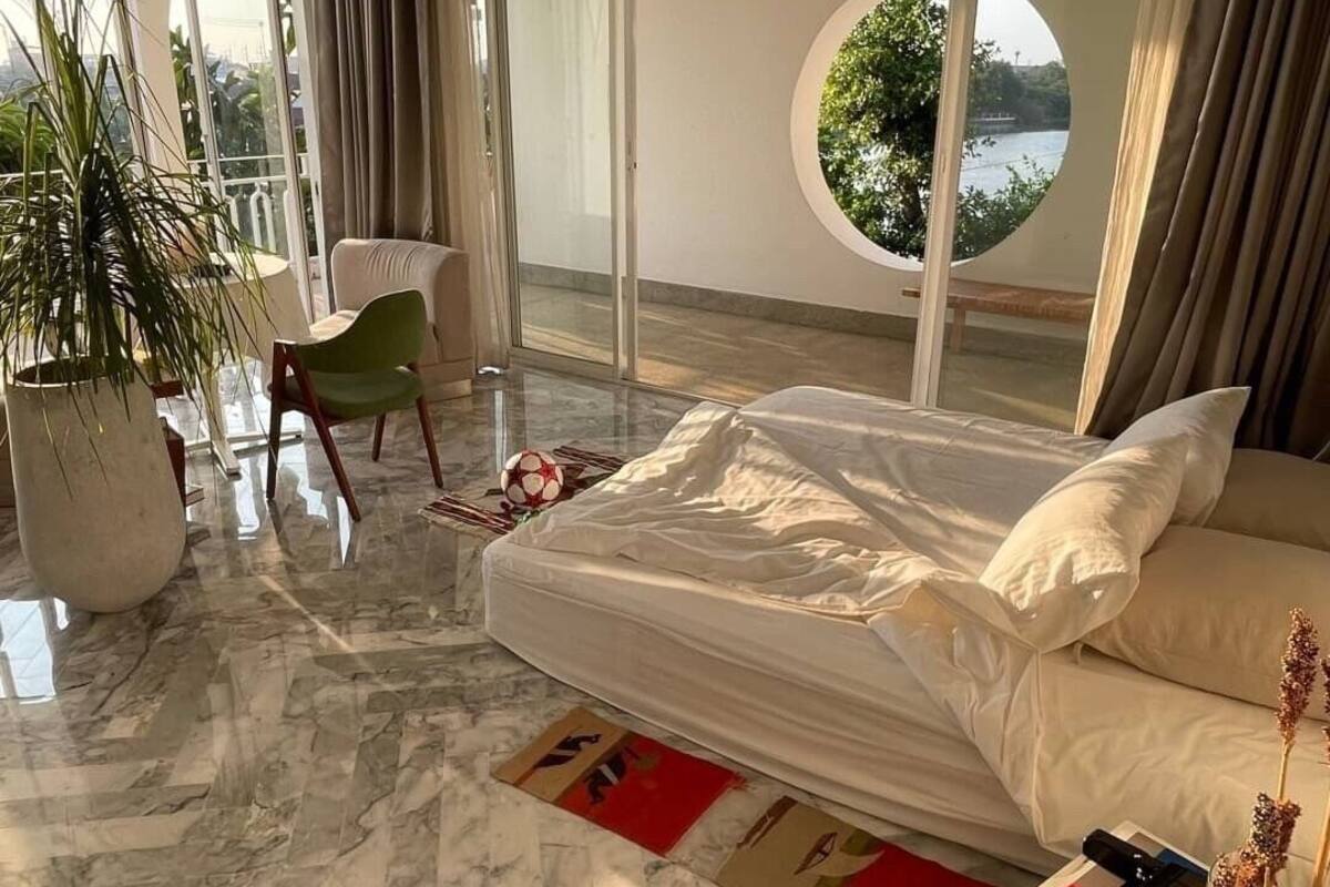 A modern bedroom with a bed, a green chair, a large potted plant, and a view of a body of water through a circular window.