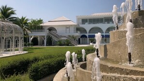 A multi-level fountain with water jets, a white building with arched windows, and a palm tree in the background. - CASA PAPA Home&Space (Nakhon Chai Si)