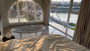 A bedroom with a large bed, a round bathtub, and a view of a bridge and water. - CASA PAPA Home&Space (Nakhon Chai Si)
