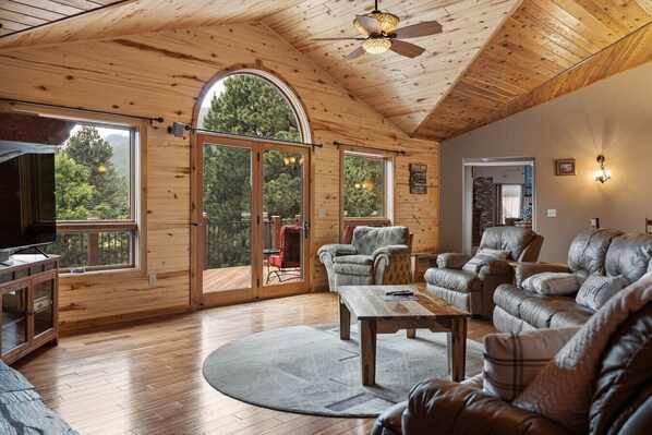 A spacious living room with wooden floors, a ceiling fan, a large arched window, and a sliding glass door leading to an outdoor deck.