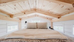 A bedroom with a sloped ceiling, a large bed, two bedside tables, and a window. - Tiny Timber- Stunning Tiny House (Spearfish)