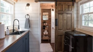 A kitchen with a wooden ceiling, a black refrigerator, and a wooden door leading to a bathroom. - Tiny Timber- Stunning Tiny House (Spearfish)