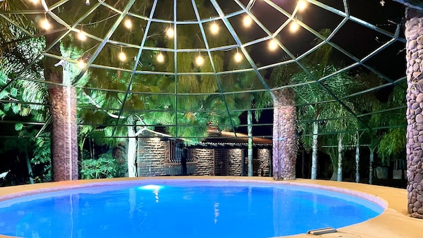 Piscina interior com superfície de água azul e límpida, cercada por uma estrutura de vidro e plantas tropicais.