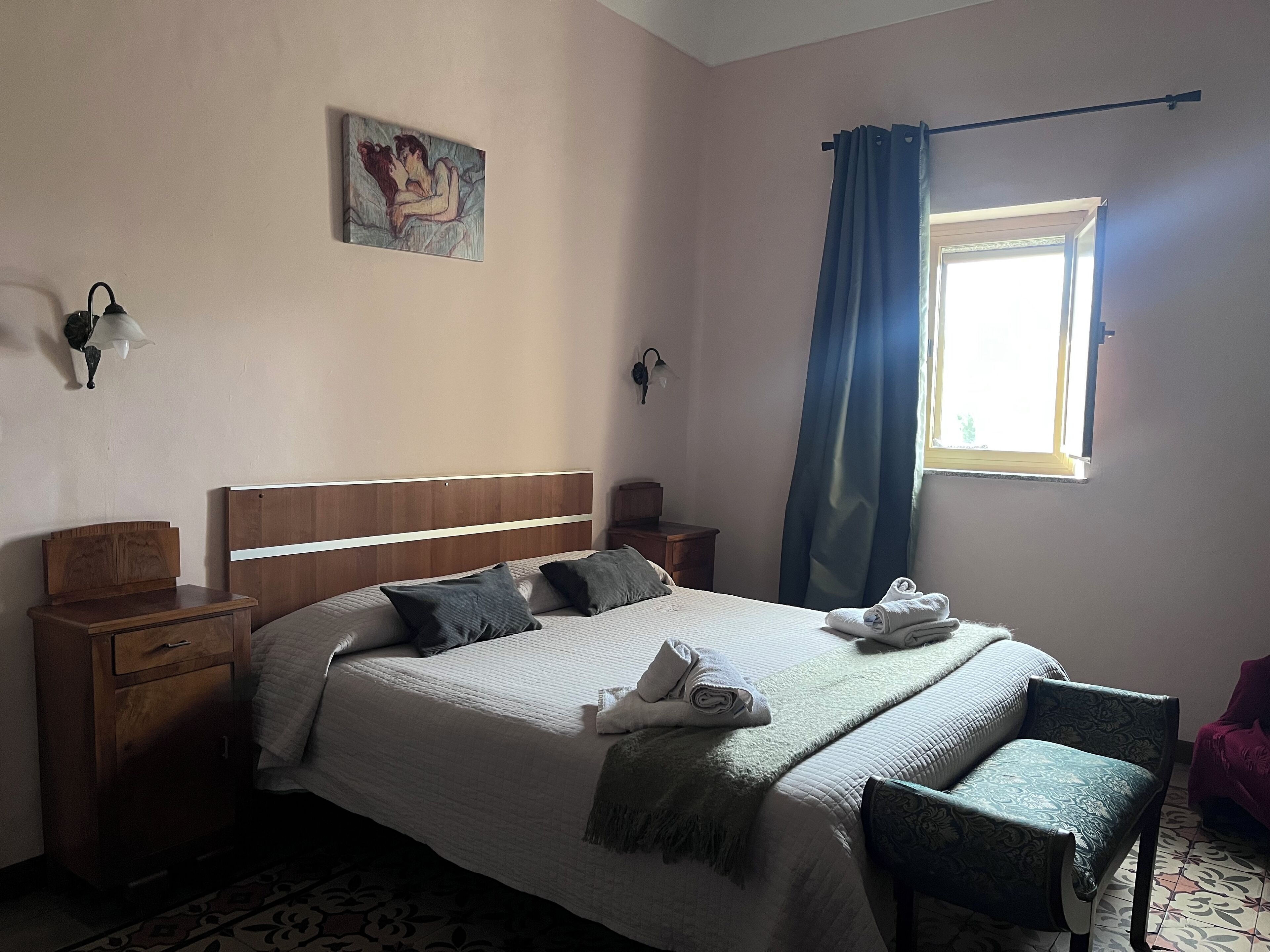 A neatly made bed with pillows, a bedside table, a chair, a window with curtains, and a wall-mounted lamp.