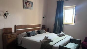 A neatly made bed with pillows, a bedside table, a chair, a window with curtains, and a wall-mounted lamp.