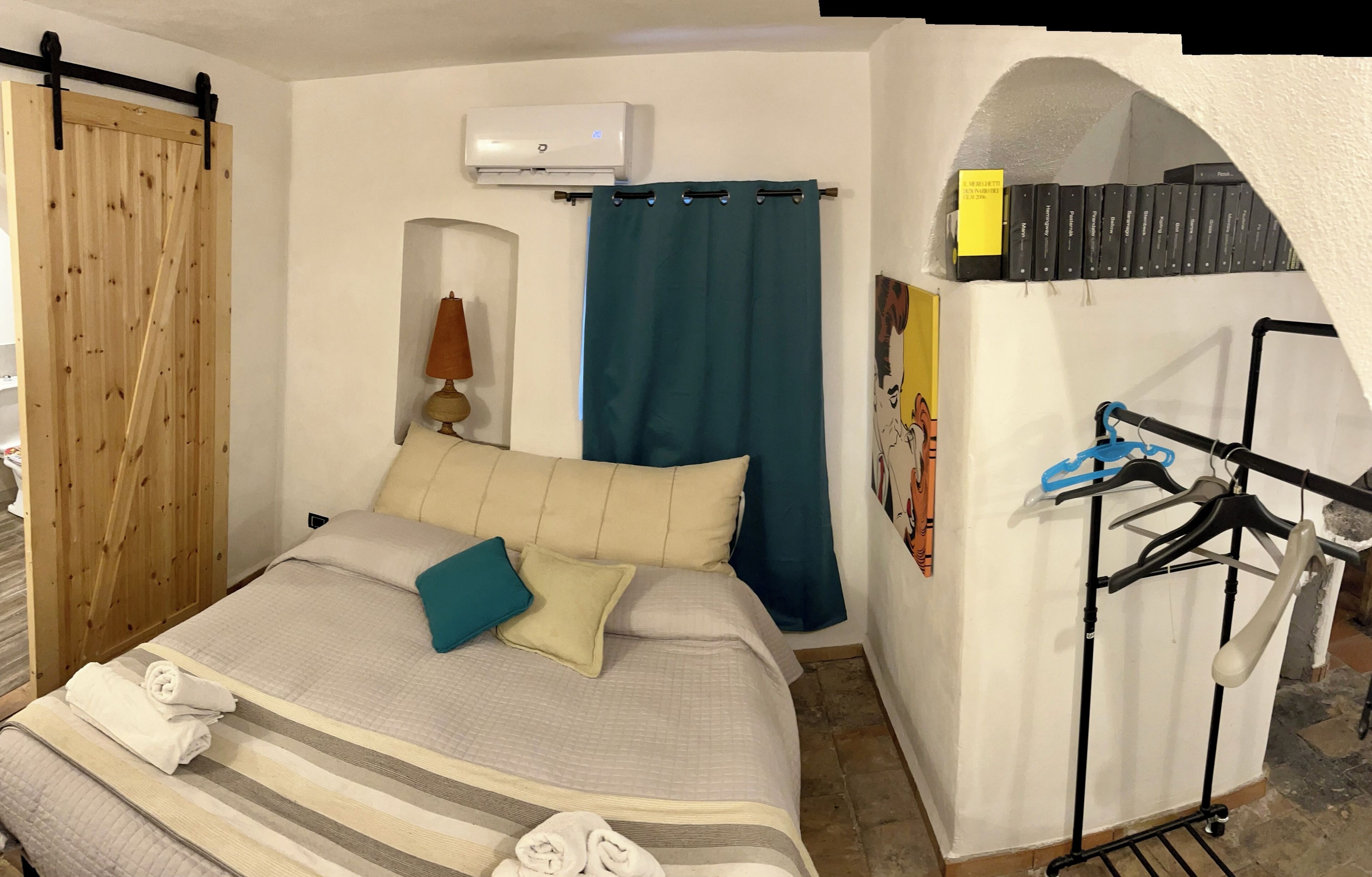 A bedroom with a bed, a wooden sliding door, a blue curtain, an air conditioning unit, a lamp, a wall-mounted shelf with books, and a clothing rack.