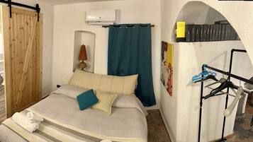 A bedroom with a bed, a wooden sliding door, a blue curtain, an air conditioning unit, a lamp, a wall-mounted shelf with books, and a clothing rack.