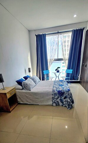 Ein modernes Schlafzimmer mit einem Bett, einem kleinen Schreibtisch und Blick auf ein Gebäude durch große Fenster.