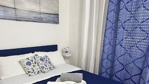A neatly made bed with a blue bedspread and white pillows, a blue and white patterned curtain, and a white headboard.