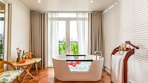 A modern bathroom with a freestanding bathtub, a wooden chair, a table with fruit and flowers, and a view of greenery through the window. - Mai Hotel  (Hanoi)