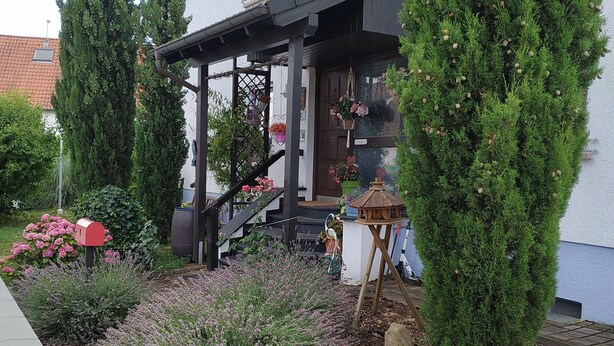 Ein weißes Gebäude mit einer überdachten Veranda, einem roten Briefkasten und einem Garten mit Lavendel und Kiefern.