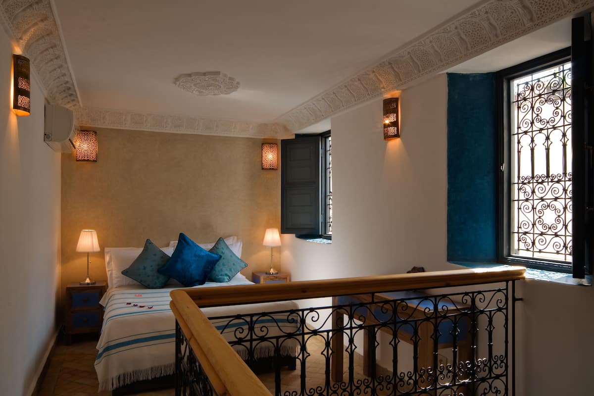 A bedroom with a bed, bedside lamps, a window with decorative iron bars, and a staircase with a wooden railing.