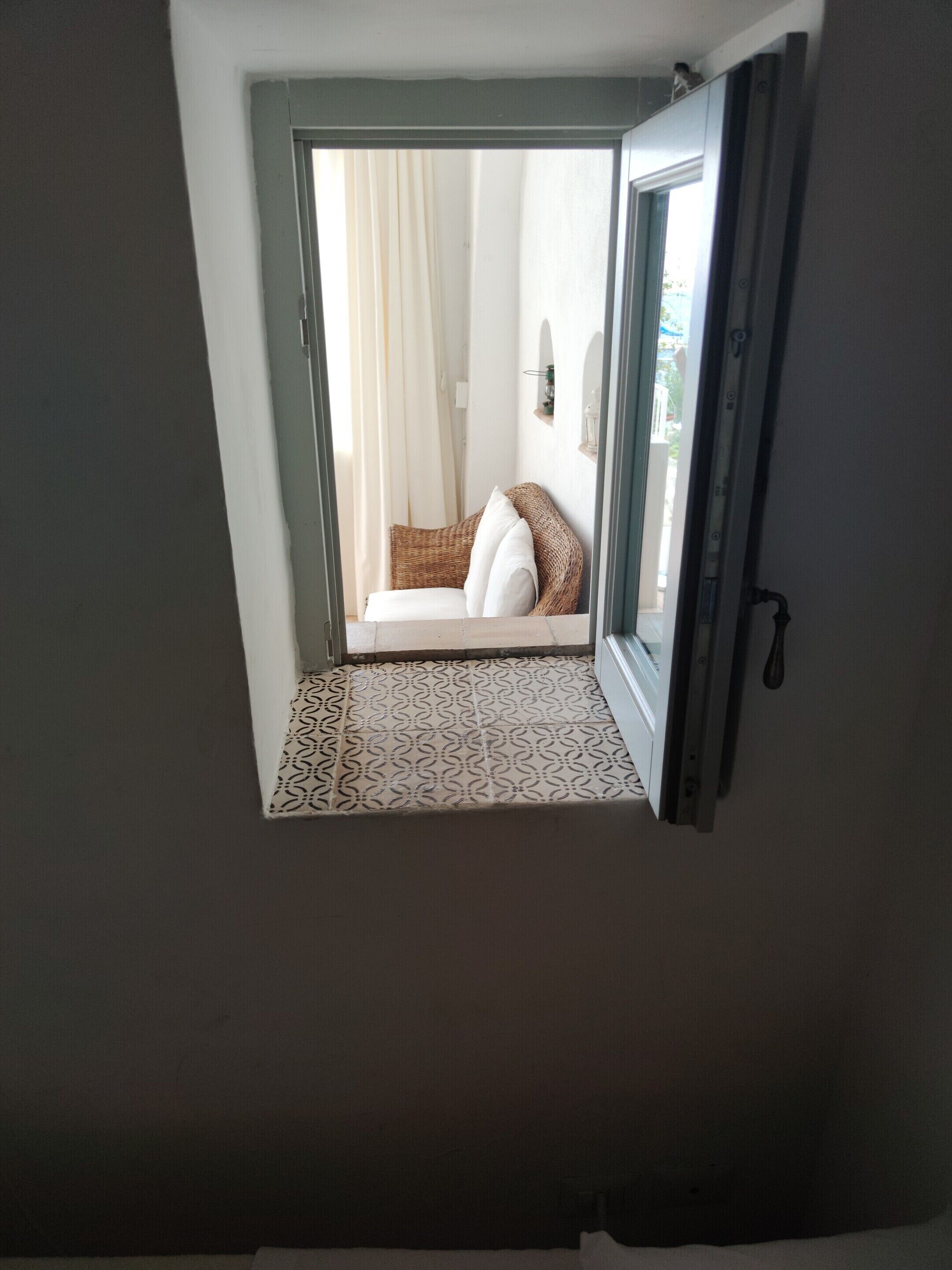 A room with a patterned floor, a wicker chair with cushions, and a view through an open door to a balcony with a white curtain.