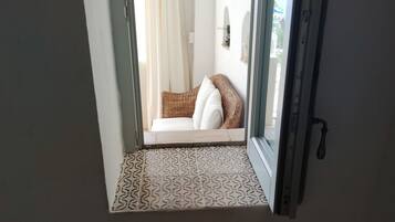 A room with a patterned floor, a wicker chair with cushions, and a view through an open door to a balcony with a white curtain.