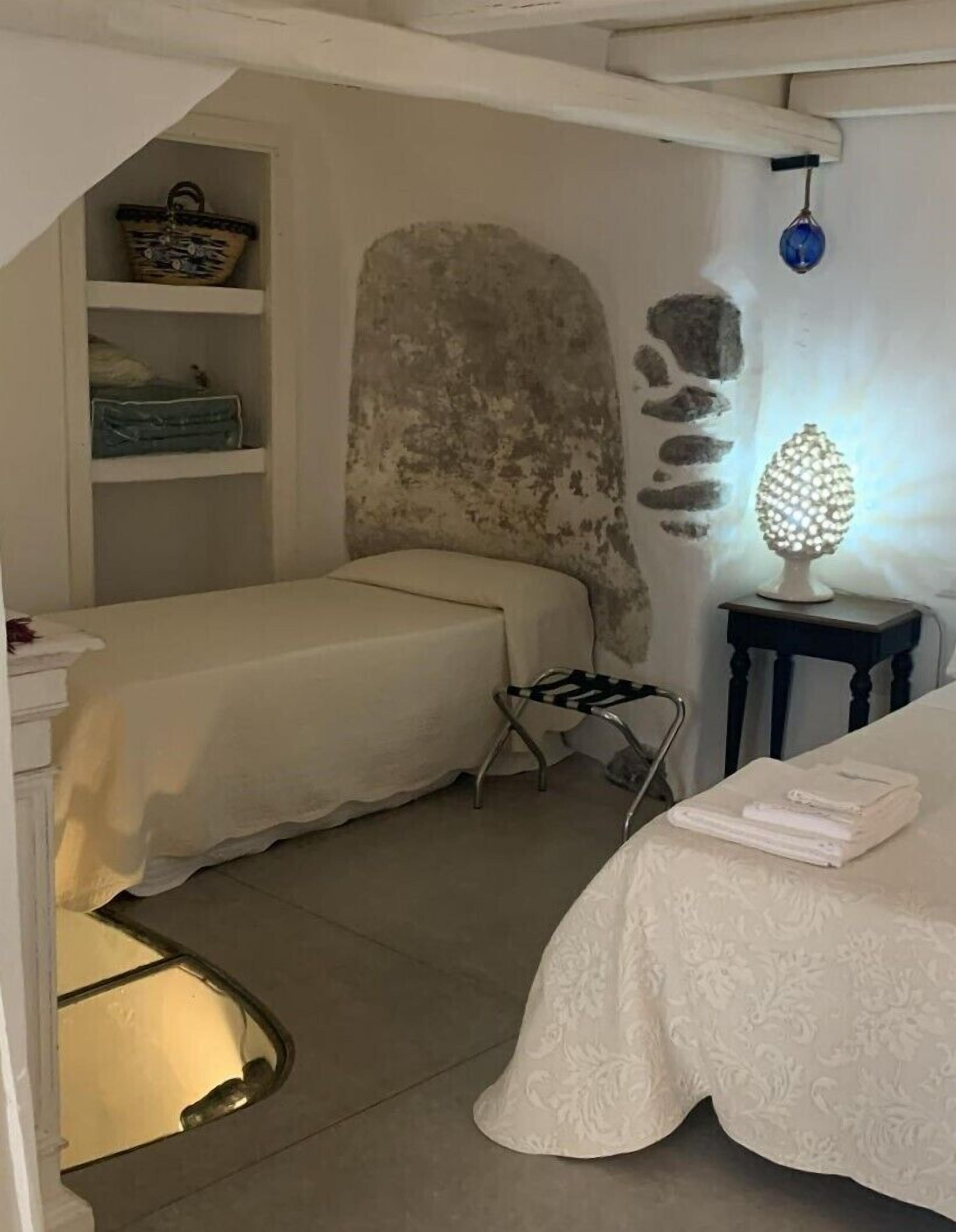 A small, cozy room with two beds, a stone wall, a shelf with a basket, and a table with a lamp.