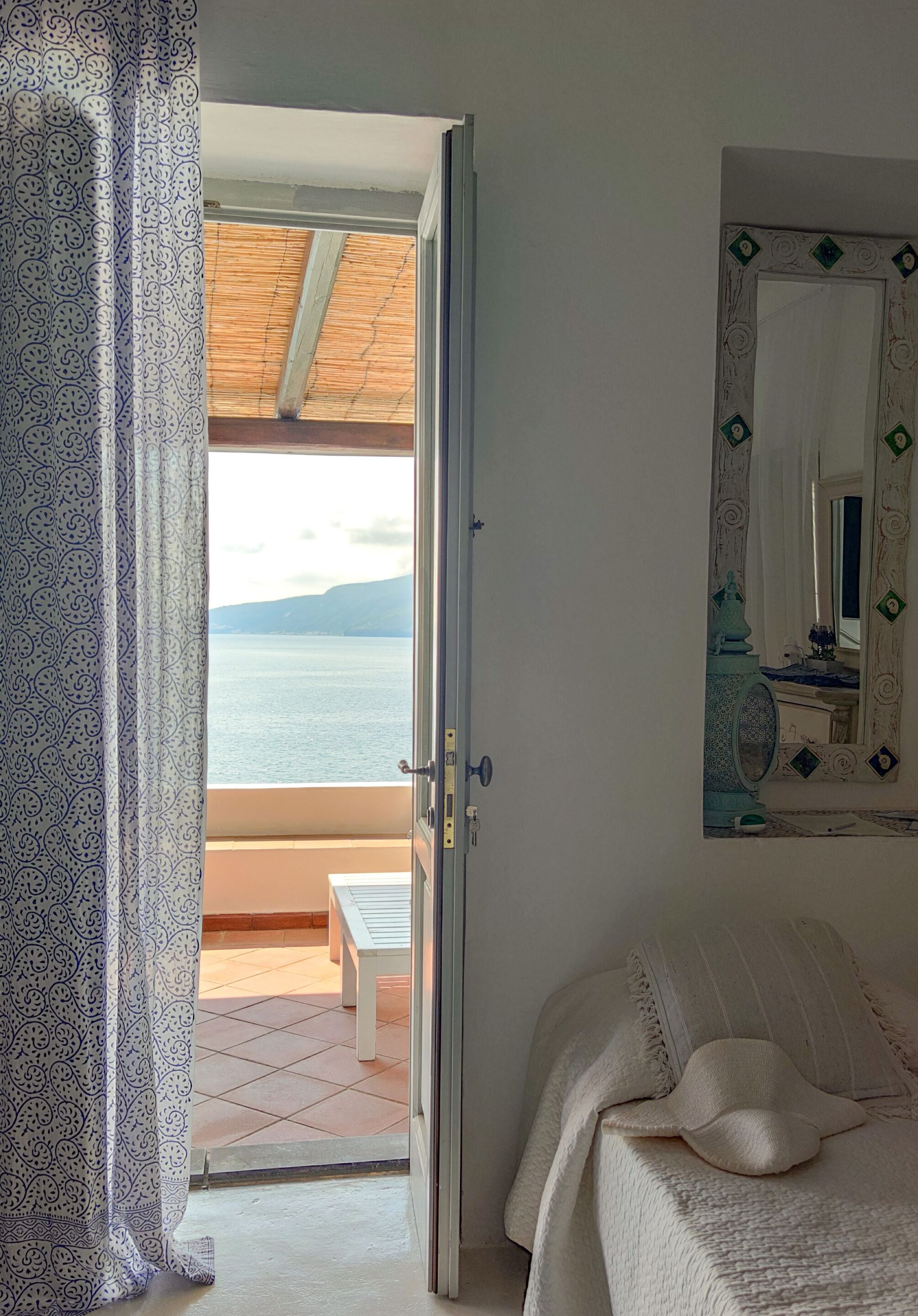 Une chambre avec un lit, une petite table et une vue sur la mer, visible par une porte ouverte.