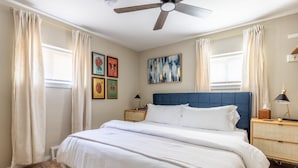 Une chambre à coucher comprenant un lit, des rideaux, un ventilateur de plafond, des tables de chevet et des œuvres d’art murales.