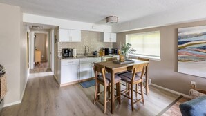 Une cuisine moderne avec des meubles blancs, une table à manger en bois et une vue sur l’extérieur par une fenêtre.