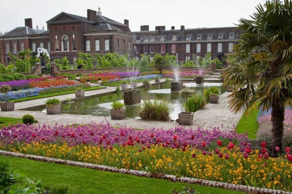 A formal garden with a fountain, a large building with a classical facade, and a statue in the foreground.