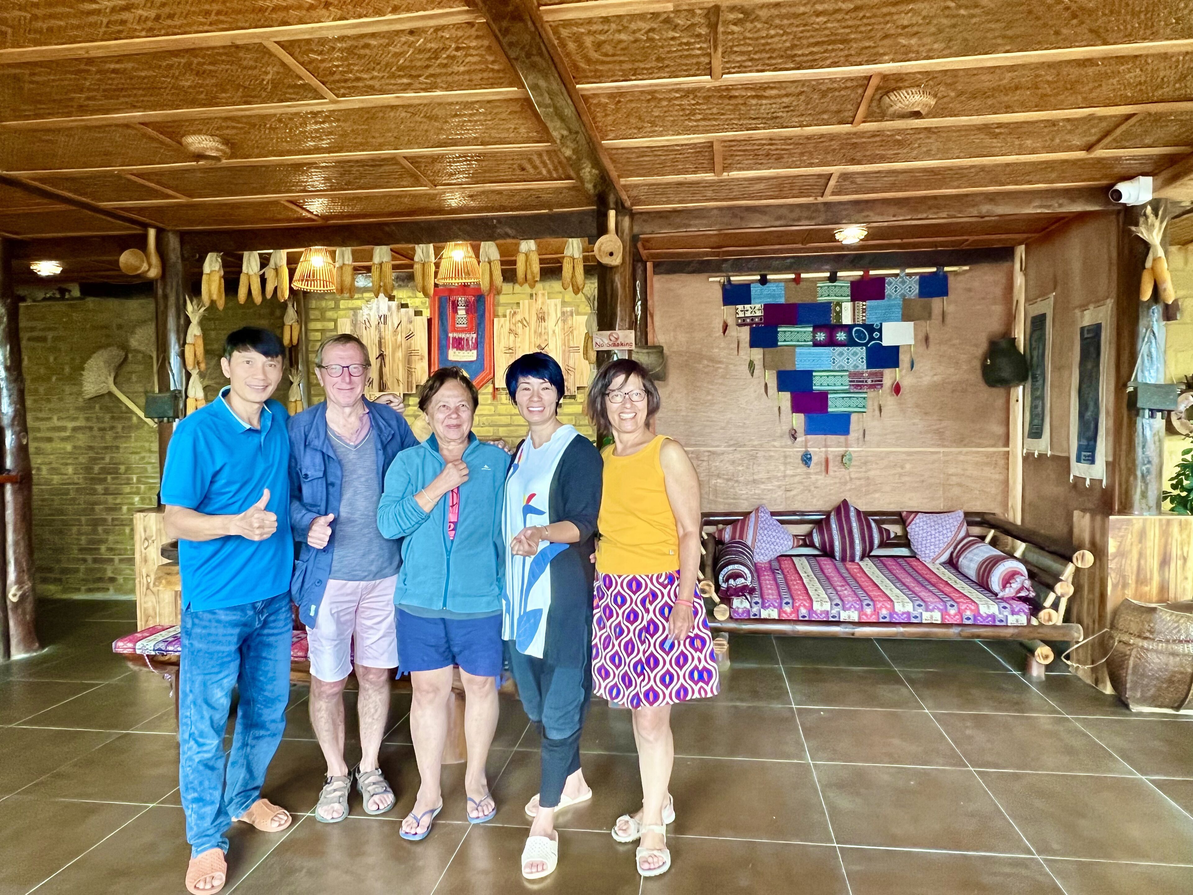 A group of people standing indoors with a wooden ceiling and a bed with patterned bedding in the background.