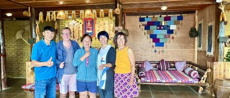 A group of people standing indoors with a wooden ceiling and a bed with patterned bedding in the background.