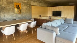 A modern living room with a wooden dining table, white chairs, a grey sofa, and a television in the background.