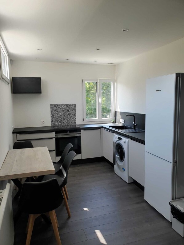 A compact kitchen with a washing machine, a refrigerator, a dining table with chairs, and a window with a view of greenery.