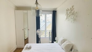 Une chambre à coucher avec un lit, une table de chevet avec une lampe, un ventilateur sur pied et une fenêtre avec des rideaux.