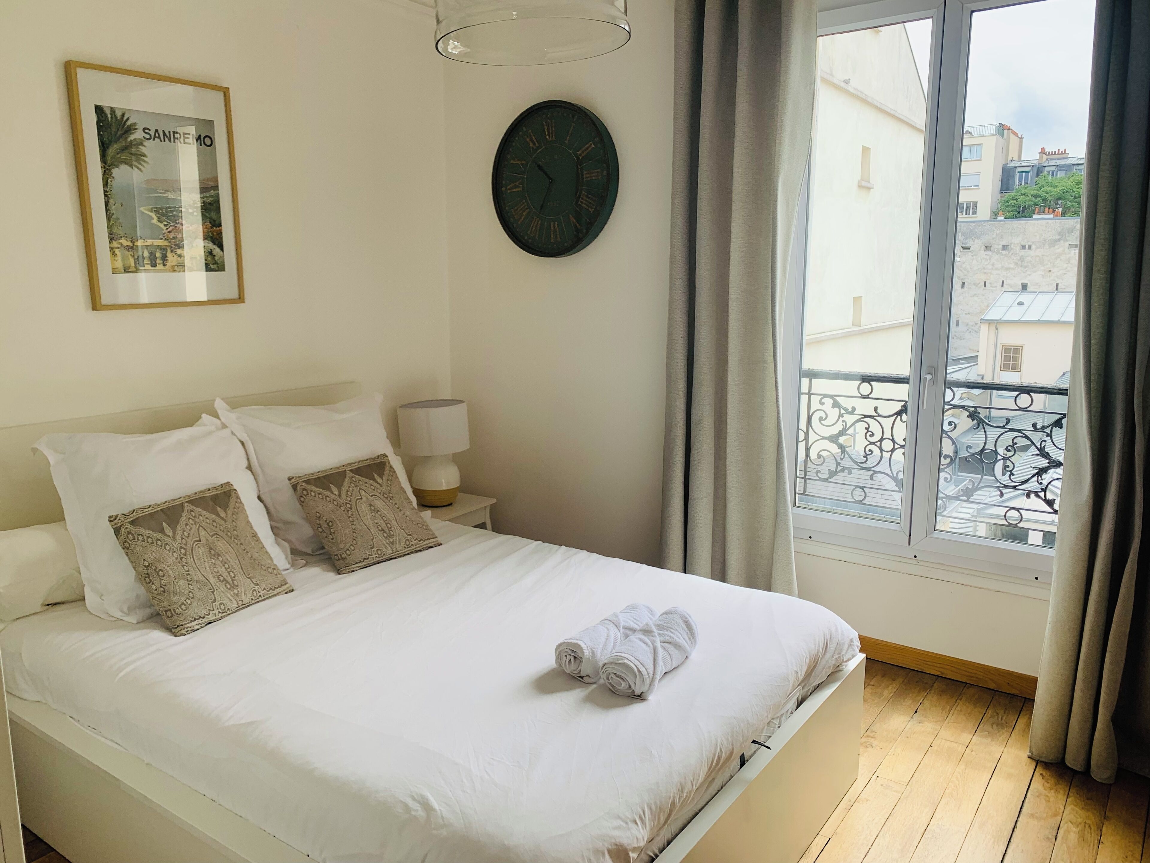 Un lit bien fait, avec du linge de lit blanc, une horloge au mur et une fenêtre avec des rideaux.