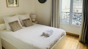 Un lit bien fait, avec du linge de lit blanc, une horloge au mur et une fenêtre avec des rideaux.