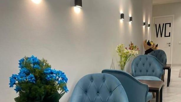 Un couloir moderne avec des chaises bleues à revêtement en tissu, une table en bois ornée d’un vase de fleurs bleues et des luminaires fixés au mur.