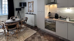 Ett modernt kök med vita skåp, en inbyggd ugn och en kaffebryggare. Det finns en matplats med flätade stolar och ett bord, samt ett stort fönster med svarta gardiner.