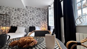 Ett hotellrum med en säng, en flätad stol, ett bord med en croissant och en mugg, och ett fönster med gardiner.