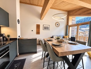 A modern kitchen with a dining area, wooden ceiling beams, and a view of a mountainous landscape.