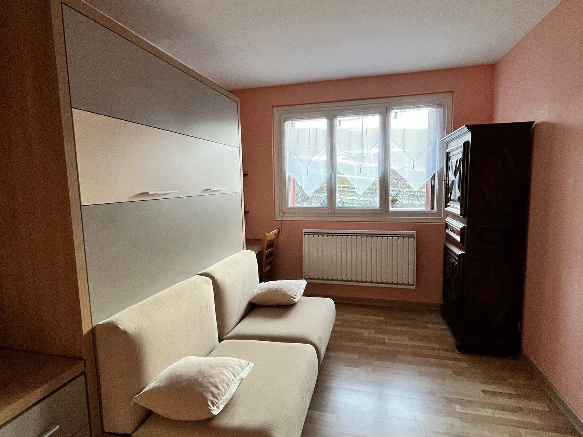 Une petite pièce agréable, avec un canapé beige, un radiateur, une fenêtre avec des rideaux et un meuble de couleur sombre.