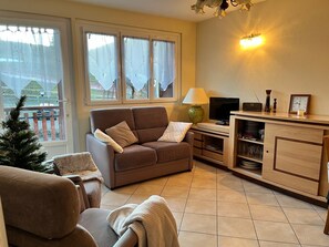 Ein gemütliches Wohnzimmer mit einer braunen Couch, einem kleinen Tisch mit Lampe, einem Fernseher und einer Schrankwand mit Regalen.