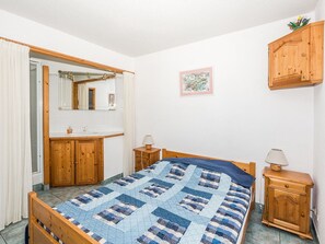 A bedroom with a wooden bed, a wooden wardrobe, a small bedside table with a lamp, a mirror above the sink, and a framed picture on the wall.