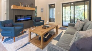 Una sala de estar moderna con chimenea, sofás azules, una mesa de centro de madera y un ventanal con vistas a árboles.