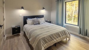 Un dormitorio con una cama, dos mesitas de noche, una lámpara de pared y un ventanal con cortinas.