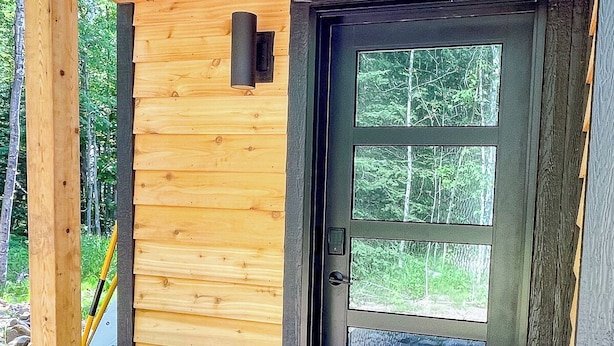 Entrada de una cabaña de madera con una puerta negra, un pequeño porche y un felpudo.