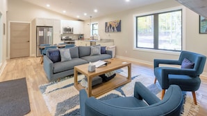 Una sala de estar moderna con un sofá gris, sillones azules, una mesa de centro de madera y una cocina al fondo.