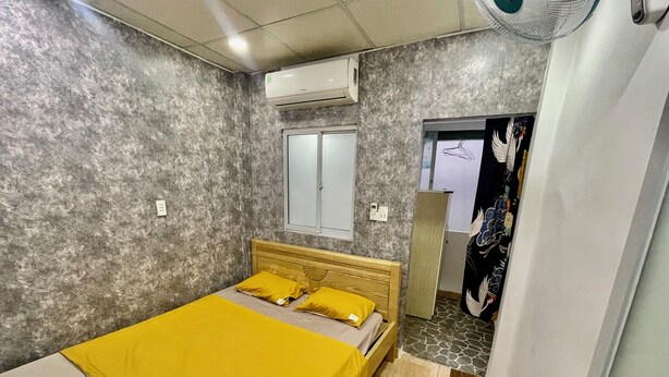 A bedroom with a bed, a window, an air conditioner, a fan, and a door with a curtain. - Enjoy Homestay  (Ho Chi Minh City)