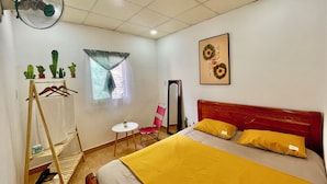 A hotel room with a bed, a standing fan, a small table, a chair, a mirror, and a window with curtains. - Enjoy Homestay  (Ho Chi Minh City)