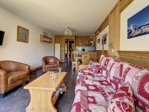 Uma sala de estar acolhedora com um sofá, uma poltrona de couro e uma mesa de centro de madeira. Ao fundo, dispõe-se uma área de cozinha com armários de madeira e uma mesa de jantar.