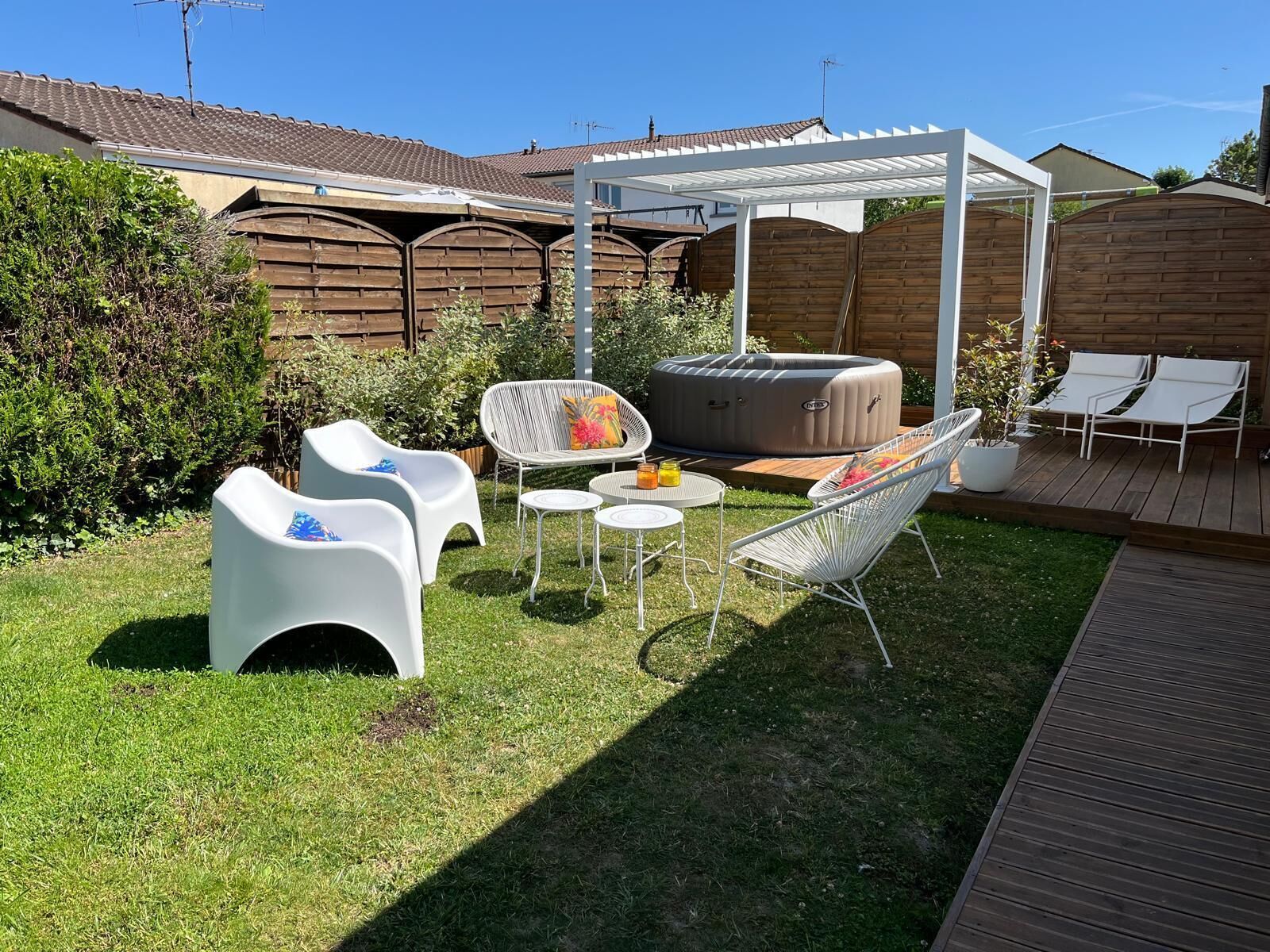A backyard with a wooden deck, a white pergola, outdoor furniture including chairs and a table, and a hot tub.