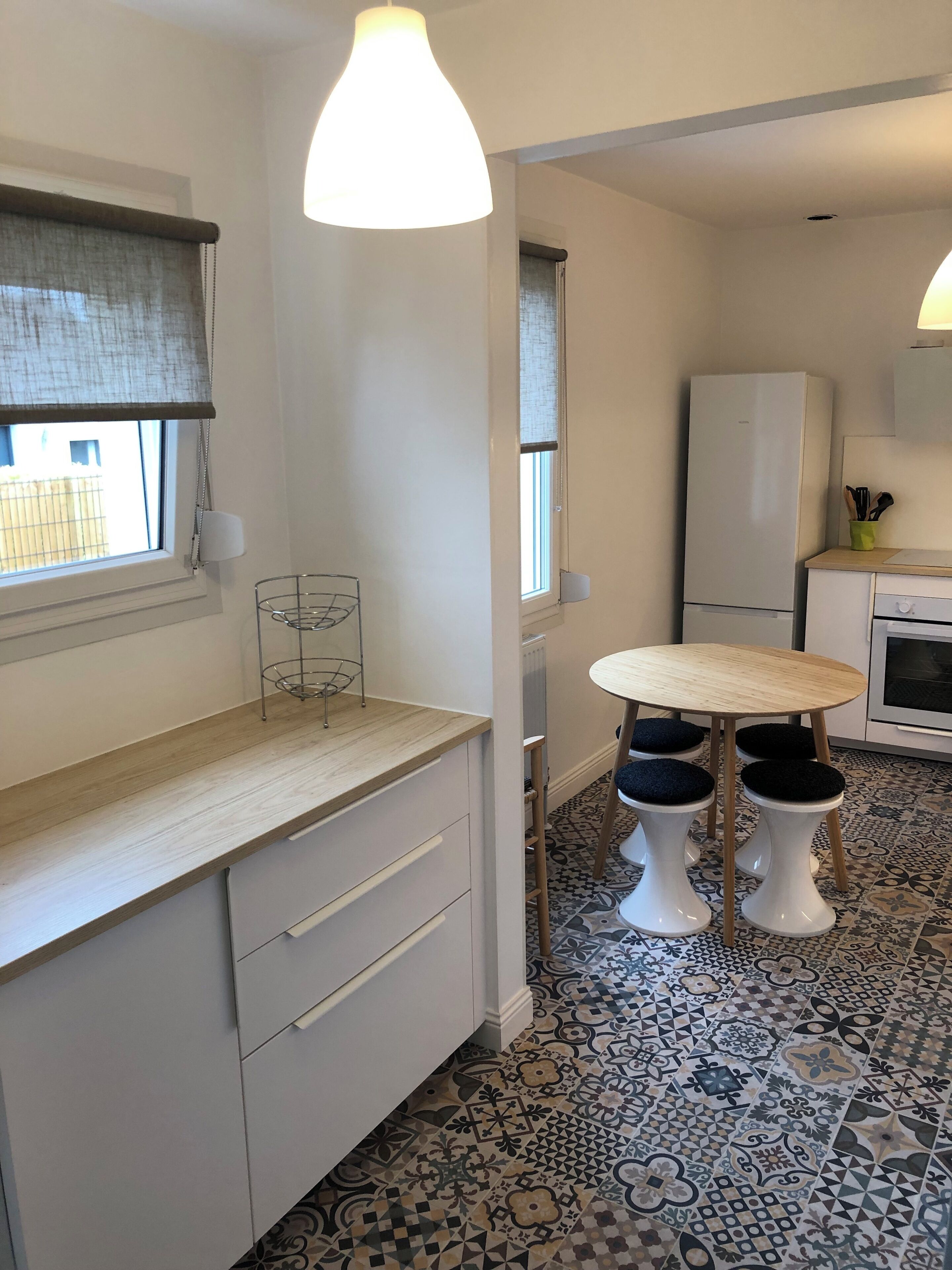A compact kitchen with a wooden countertop, a round table with stools, a white refrigerator, and patterned floor tiles.