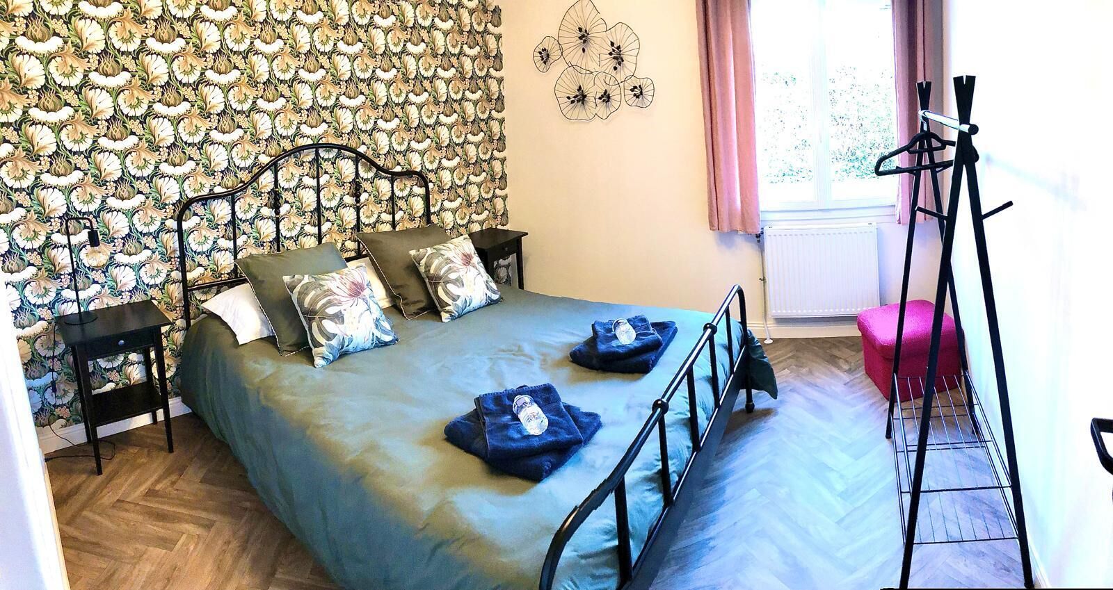 Un dormitorio con papel tapiz floral, una cama con cubrecama azul, dos mesitas de noche, una ventana con cortinas y un trípode.