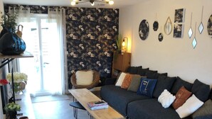 En moderne stue med en sofa, et sofabord og en stilet stol. Der er et bogreol med dekorative genstande og et stort vindue med gardiner.