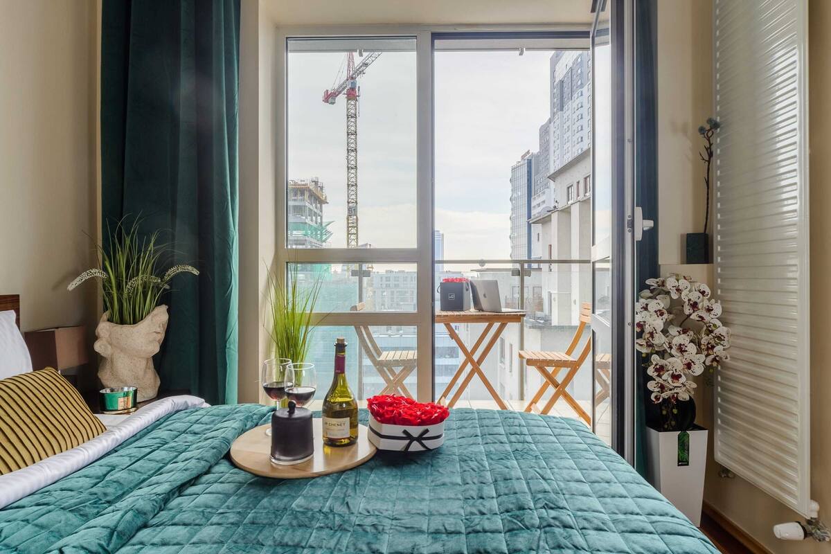 Quarto de hotel com cama, bandeja com bebidas, vaso de flores e vista para uma grua e prédios.