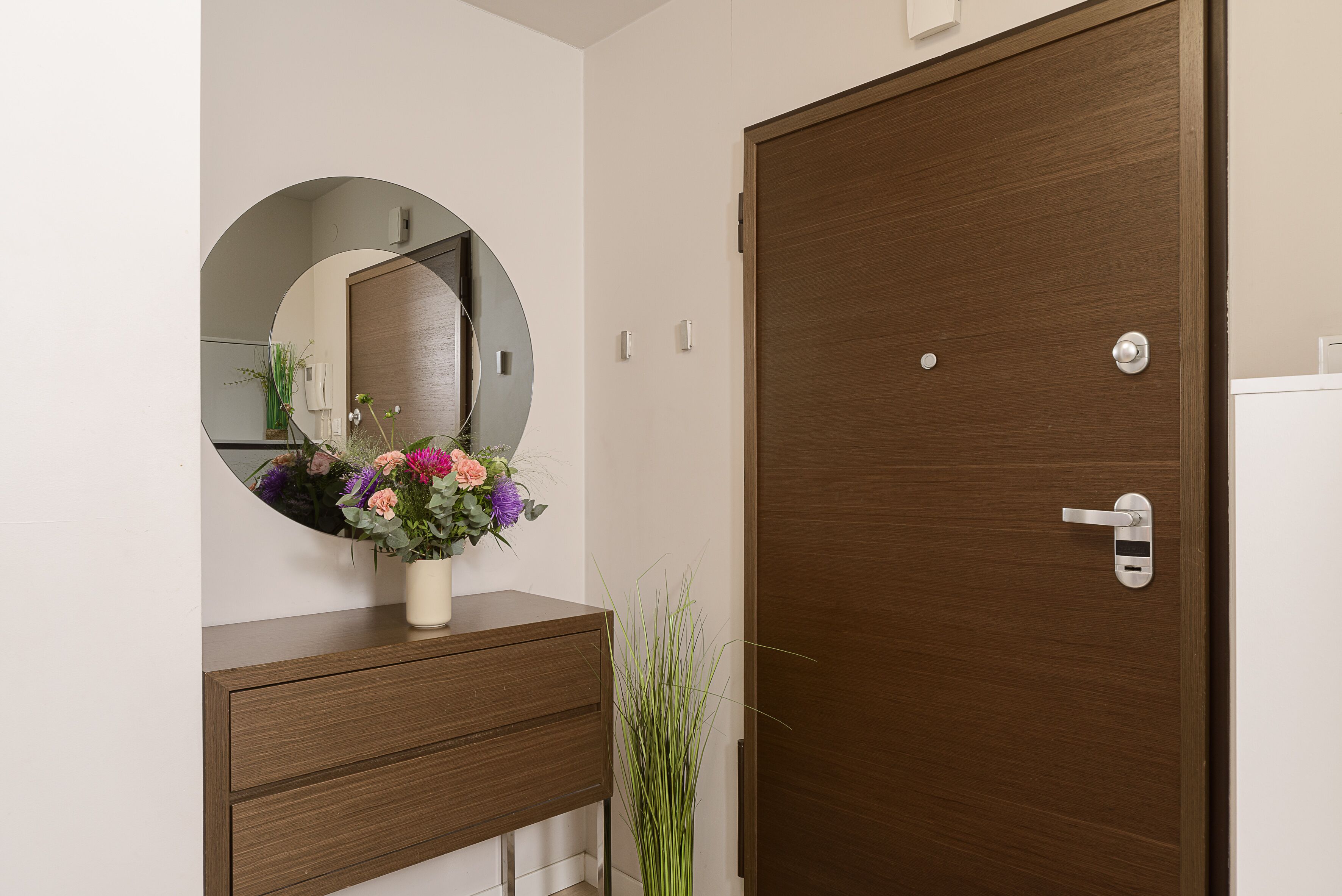 Eine Holztür mit modernem Griff, ein runder Spiegel über einer Holzkommode und eine Vase mit Blumen.